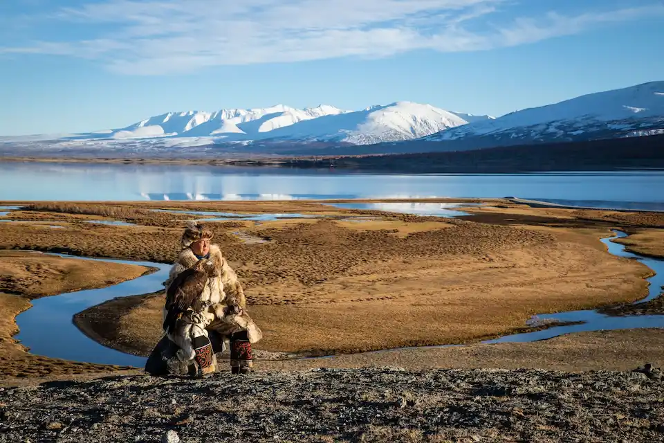 A Kazakh eagle hunter rests on a rock in western Mongolia, his golden eagle perched proudly against the backdrop of the Altai Mountains
