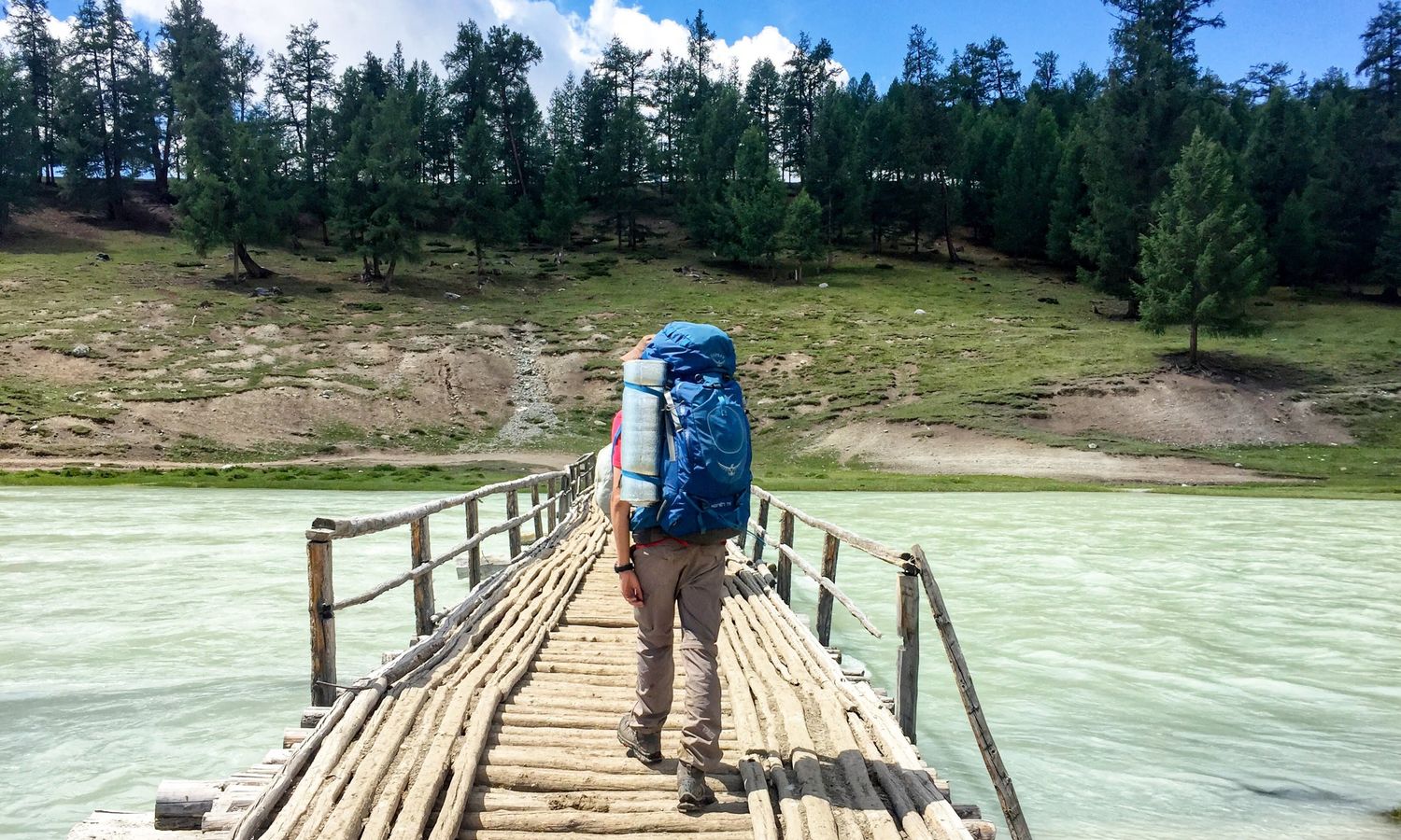 A trekker with a large backpack crossing a wooden log bridge over a glacial river
