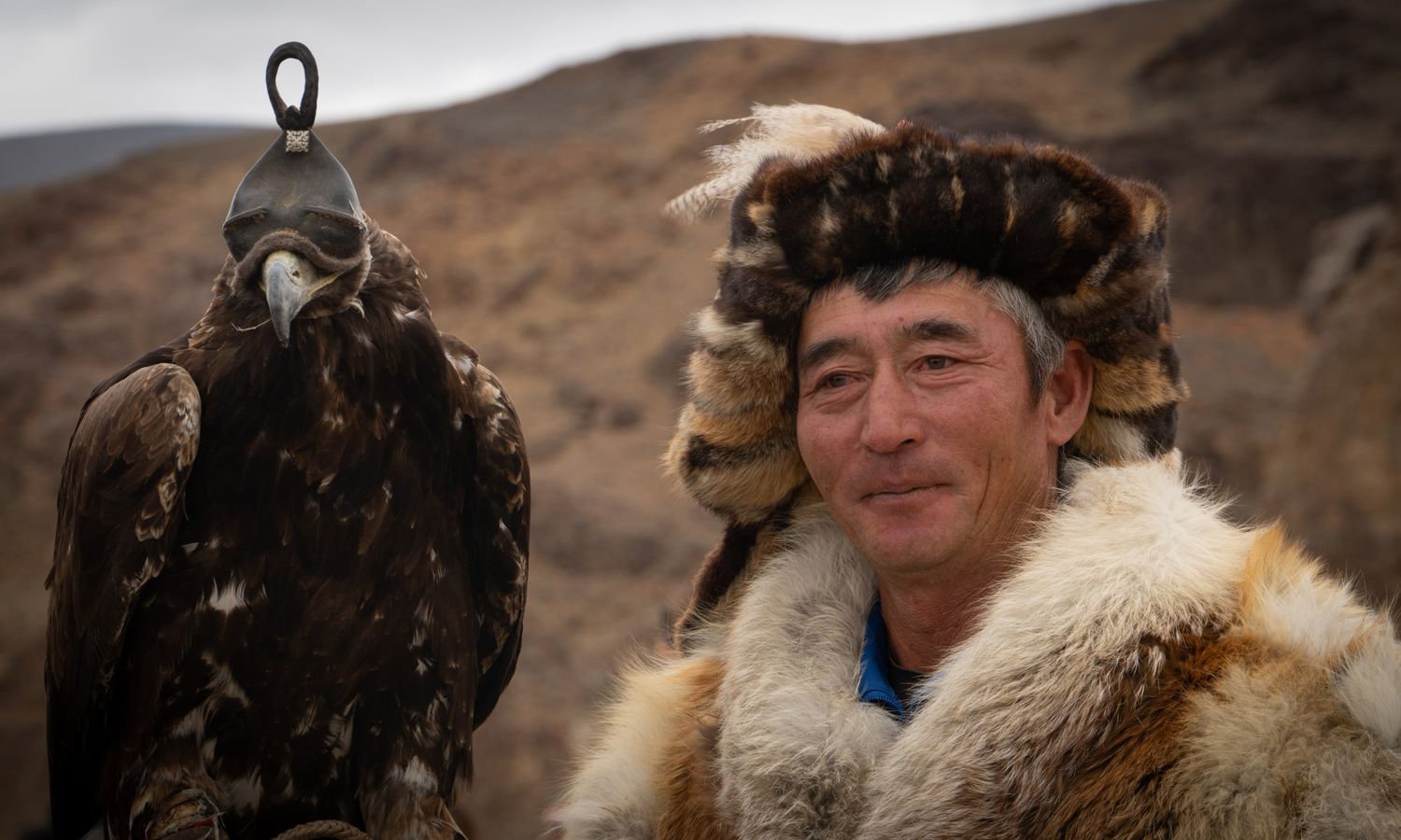 A smiling Kazakh eagle hunter in a fox-fur hat beside his hooded golden eagle